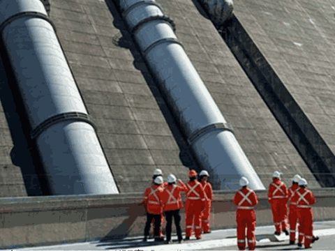 A group of workers clad in orange overalls and hard hats, standing on the concrete dam at the bottom of several very large pipes laid against the slope of the Revelstoke dam.
